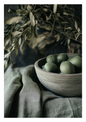 A rustic ceramic bowl filled with green olives, set on draped olive-green linen fabric with dried olive branches in the background.